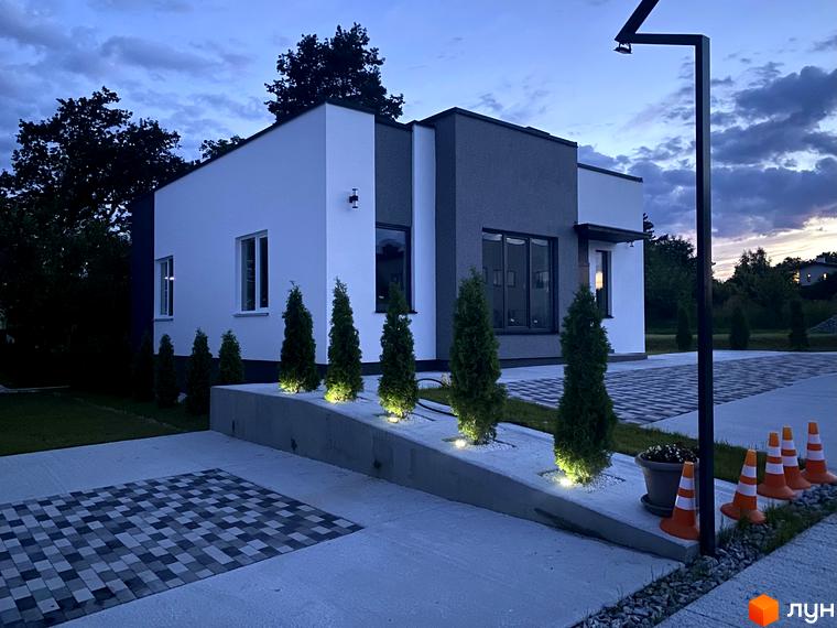 Сучасний двоповерховий будинок з білою та сірою обробкою, розташований на доглянутій прибудинковій території з туями та декоративним освітленням.