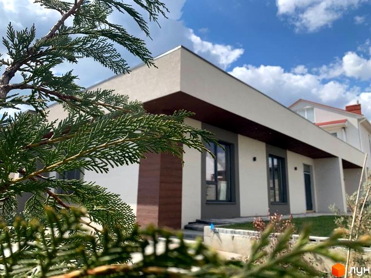 Зображення будинку в Котеджному містечку La Vida. Двоповерховий будинок з білими стінами та коричневими вставками, видно зелений газон та дерева.