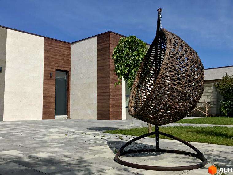 Вигляд Котеджного містечка La Vida з плетеним коконом-гойдалкою на подвір'ї. Двоповерховий будинок з оздобленням під дерево та камінь.