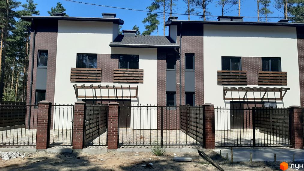 Зображення Котеджного містечка Лісове-2: двоповерхові будинки з цегляним фасадом та білими вставками, огороджені кованим парканом. На території ростуть дерева.