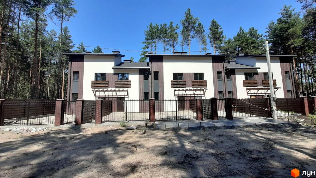 Зовнішній вигляд Котеджного містечка Лісове-2: три двоповерхові будинки з коричневою цеглою та білими стінами, огороджені парканом. Навколо сосновий ліс та піщаний ґрунт.