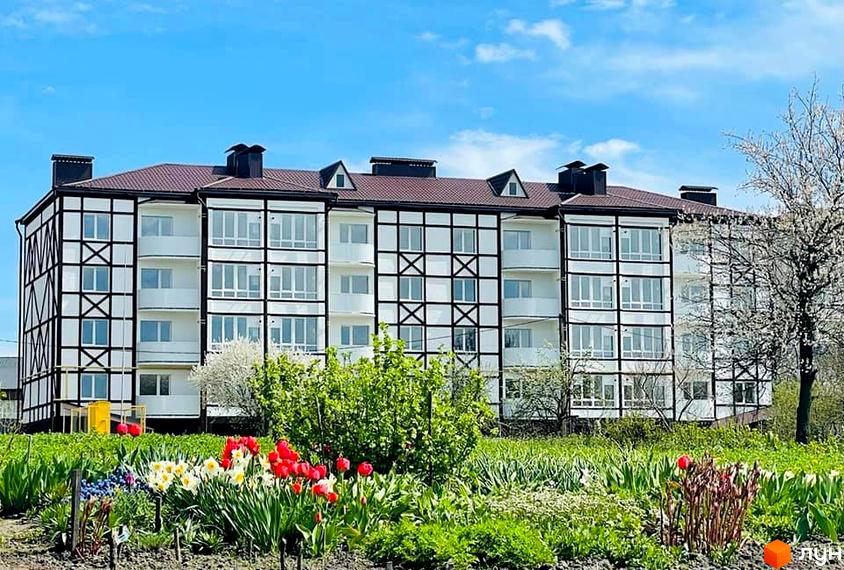 Зовнішній вигляд триповерхового будинку з коричневим дахом та білим фасадом з чорними декоративними елементами. Перед будинком розташований квітник з тюльпанами та нарцисами.