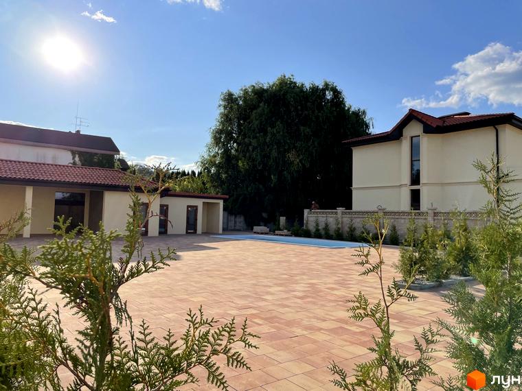 Котеджне містечко КМ Фонтан: двоповерховий будинок з червоним дахом, велика вимощена тераса та басейн. Навколо зелені насадження та блакитне небо.