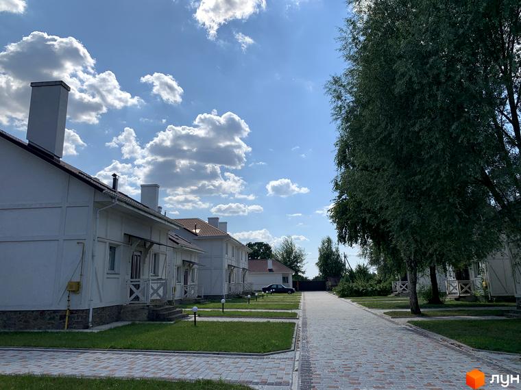 Вигляд котеджів у Котеджному містечку „Северинівська Перлина“ з акуратними газонами та брукованою дорогою. Будинки з білими стінами та димарями на тлі блакитного неба.