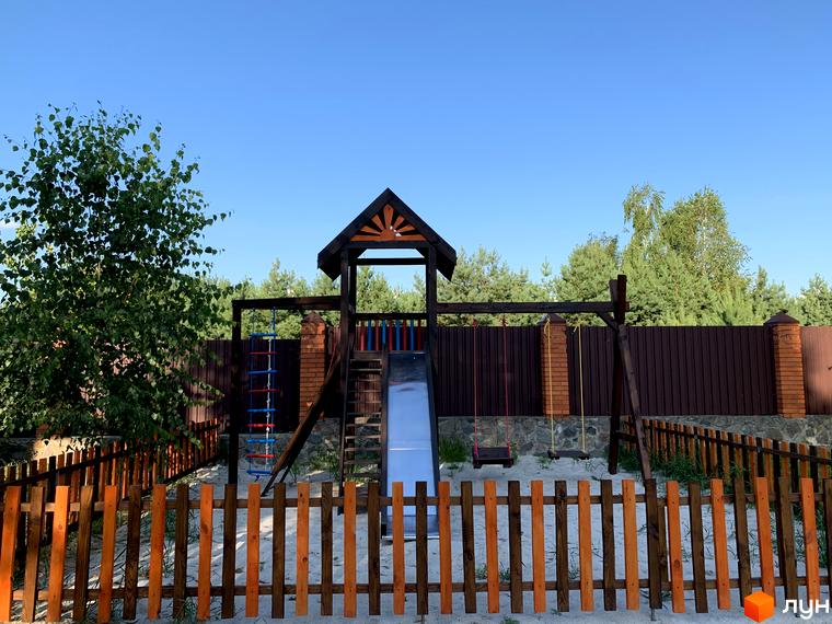 Зображення дитячого майданчика в Котеджному містечку „Северинівська Перлина“. На фото гірка, гойдалки, драбина та пісочниця, огороджені деревʼяним парканом.