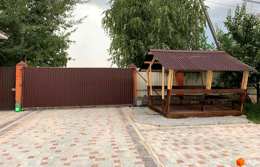 Зображення будинку в Котеджному містечку „Северинівська Перлина“. На фото видно огорожу, двір з бруківкою та альтанку.