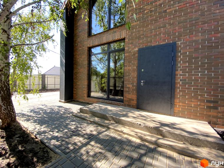 Зовнішній вигляд будинку з цегляною кладкою та великими вікнами. Прибудинкова територія з бруківкою та деревами.