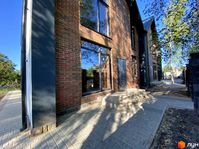 Фасад будинку з цегли та темними вставками, поруч доріжка з бруківки та зелена зона. Дуплекси Тут – ваш новий дім.