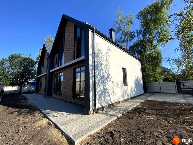 Фасад дуплексу з темно-коричневої цегли та світлої штукатурки, великі вікна, темний дах. Прибудинкова територія має вимощену доріжку та під'їзд.