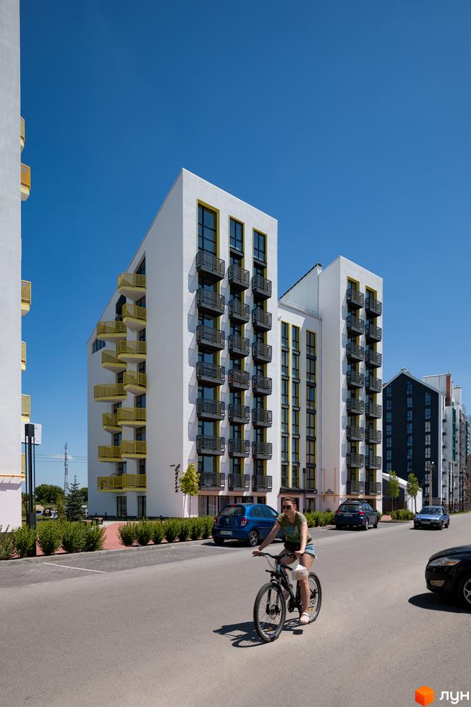 Житловий комплекс City Lake: білі багатоповерхові будинки з жовтими акцентами та темними балконами. На передньому плані дорога, зелені насадження та велосипедист.