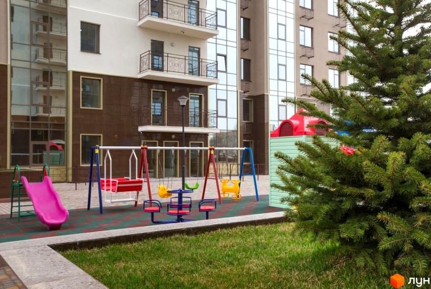 Житловий комплекс Сімнадцята Перлина. Багатоповерховий будинок з балконами та великими вікнами. На території дитячий майданчик з гіркою та гойдалками, зелена трава.