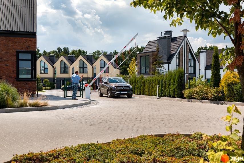 Котеджне містечко KМ VILLAGGO: фасади будинків з великими вікнами та двосхилими дахами, огороджена територія з шлагбаумом та автомобілем біля в'їзду.