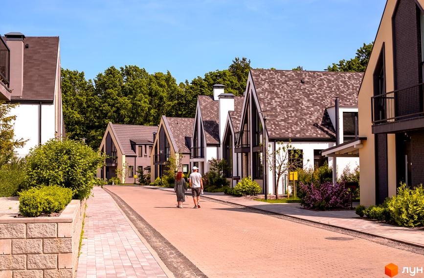 Котеджне містечко VILLAGGO. Сучасні будинки з двосхилими дахами та великими вікнами розташовані на брукованій вулиці з доглянутими зеленими насадженнями.