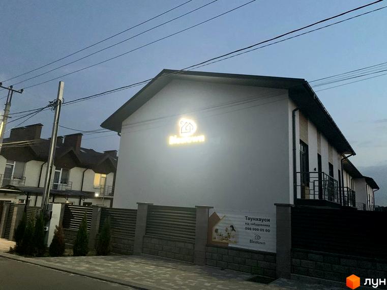 Фасад будинку Blestown з підсвіченим логотипом та огородженою територією з банером. Зовнішній вигляд новобудови та прибудинкової території.