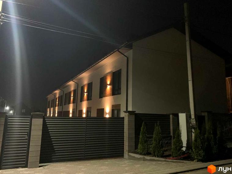 Зовнішній вигляд таунхаусів Blestown з підсвіченими вікнами та огорожею. Територія перед будинками прикрашена зеленими туями.