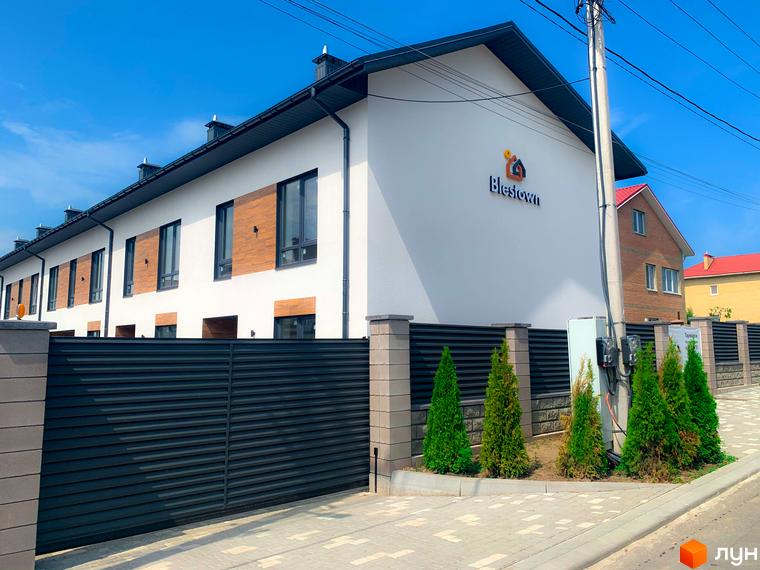 Таунхаус Blestown з білою фасадною стіною, дерев'яними вставками та темним дахом. Огороджена територія з туями та парканом.