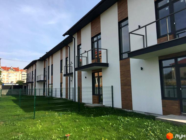 Таунхаус Blestown – це сучасна новобудова з білою та дерев'яною обшивкою фасаду та чорними балконами. Перед будинком розташований зелений газон та паркан.