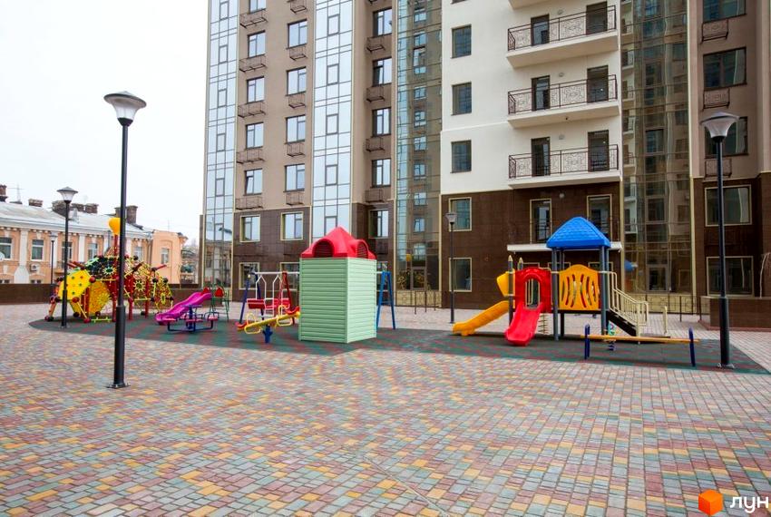 Високий житловий будинок з великими вікнами. На прибудинковій території розташований дитячий майданчик з гірками та гойдалками, а також бруківка.