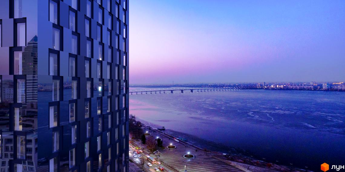 Висотна будівля з скляним фасадом на фоні річки та мосту. На передньому плані розташована набережна з освітленням та автомобілями.