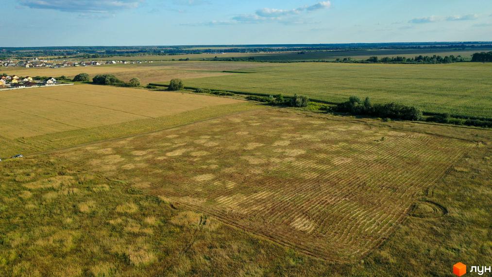 Котеджне містечко Луговий: вид з повітря на зелені поля та житлові будинки. Навколо комплексу розташовані поля з вибірково зібраною рослинністю та зелені насадження.