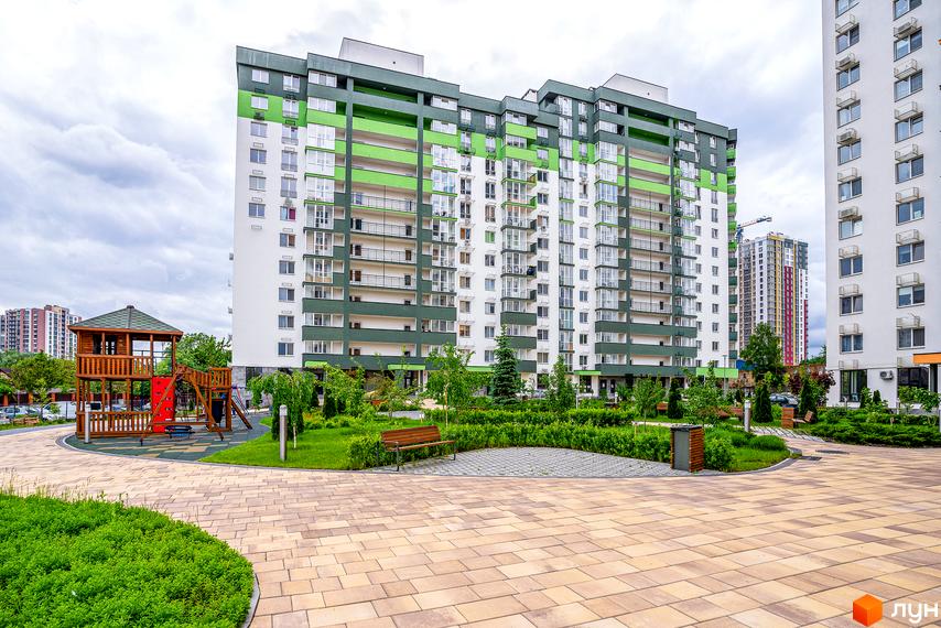 Житловий комплекс Сирецькі сади. Зелено-білий будинок з балконами та дитячим майданчиком. Територія з лавками та зеленими насадженнями.