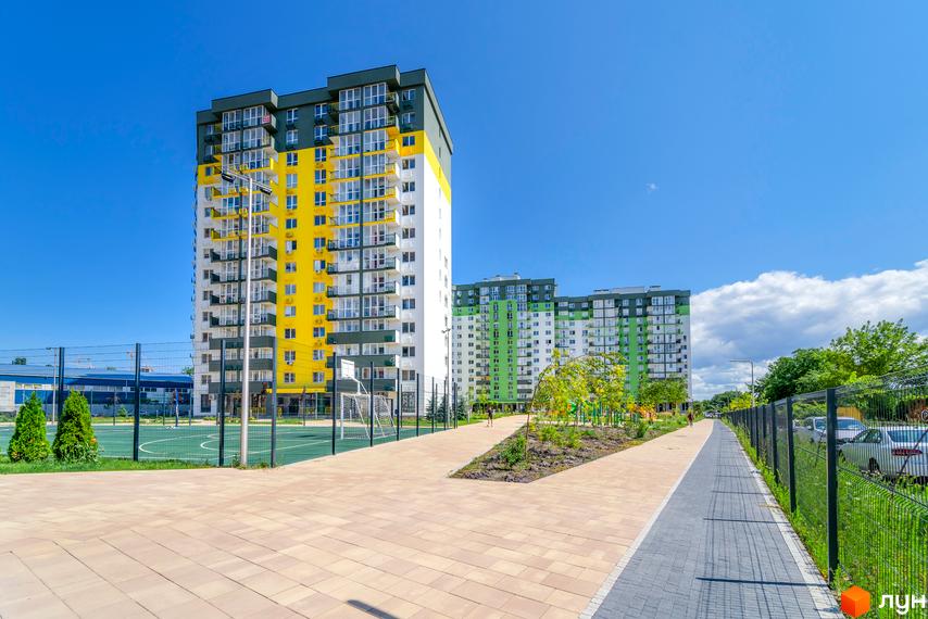 Житловий комплекс Сирецькі сади з яскравим жовто-білим фасадом та зеленими сусідніми будинками. На території облаштований спортивний майданчик та пішохідні доріжки.