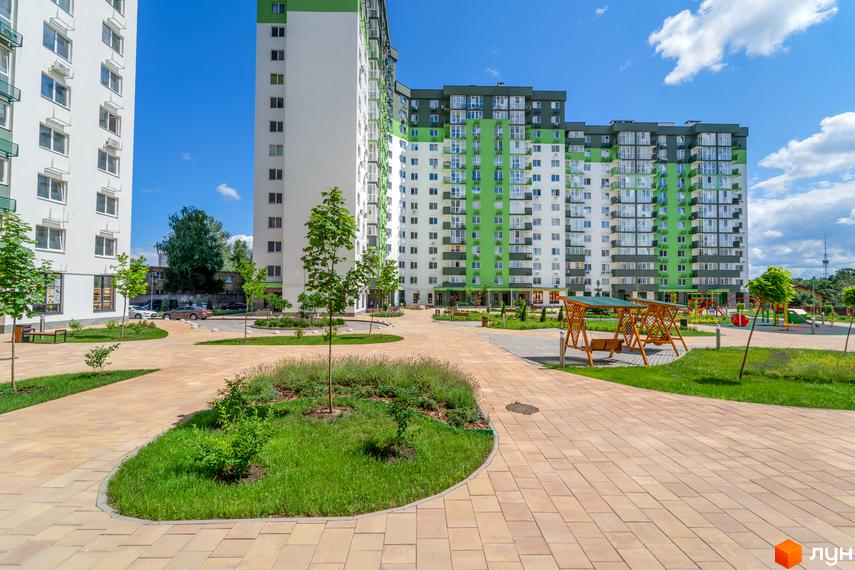 Зовнішній вигляд новобудови ЖК Сирецькі сади з білими та зеленими фасадами. Прибудинкова територія з бруківкою, газоном та дитячим майданчиком.