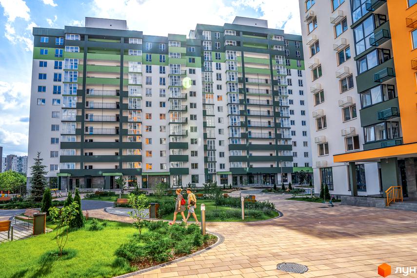 Світлий житловий комплекс Сирецькі сади з зеленими та білими фасадами, балконами та кондиціонерами. На прибудинковій території зелений газон, дерева та лавки.