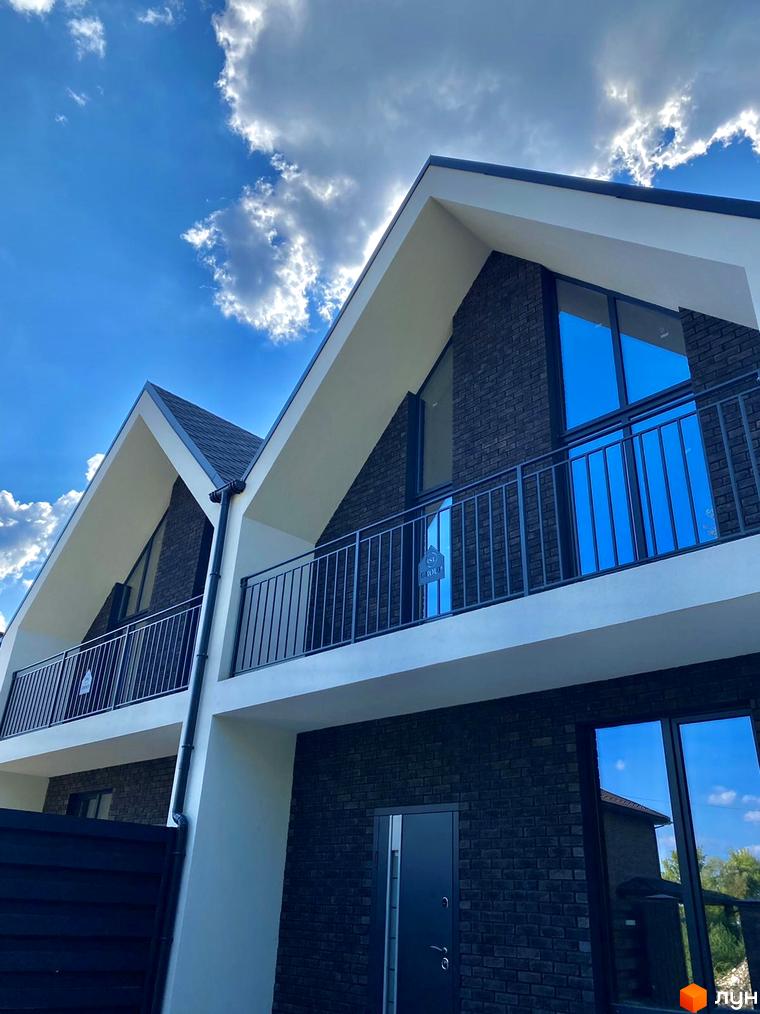 Дуплекс MANHATTAN з цегляним фасадом та великими вікнами під блакитним небом з хмарами. Навколо будинку видно елементи прибудинкової території.