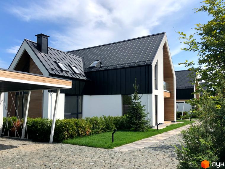 Котеджне містечко КМ Hill Park з чорними металевими дахами та білими стінами, оточене зеленню та вимощеною доріжкою.