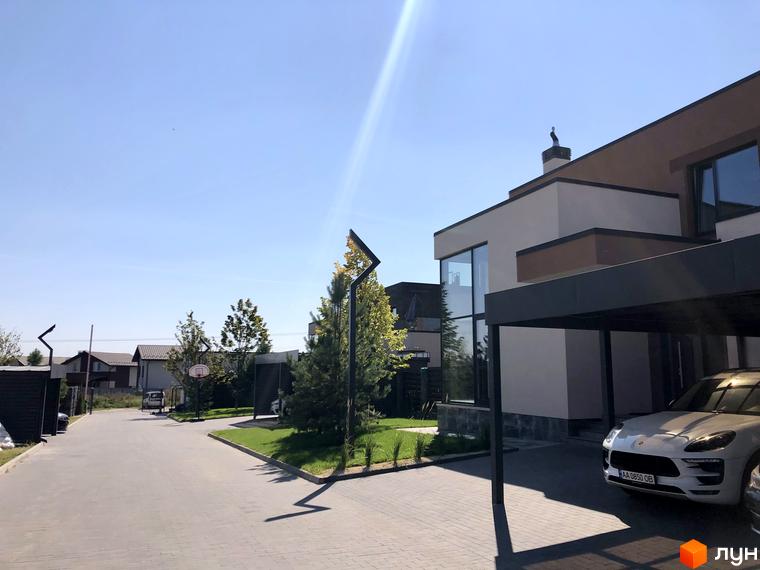 Будинок з великими вікнами та навісом для автомобіля в Містечку котеджів Crystal Forest. На території є зелені насадження та дорога з бруківки.