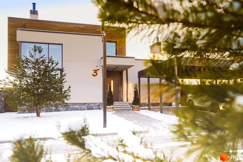Зимовий вигляд будинку з великими вікнами та дерев'яним оздобленням. Біля будинку засніжена прибудинкова територія та дерева.
