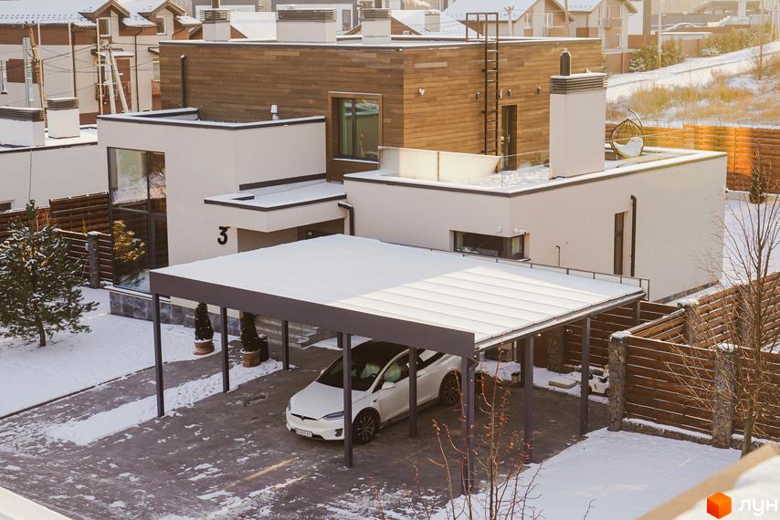 Котеджне містечко Crystal Forest. Двоповерховий будинок з білими стінами та дерев'яним оздобленням. Перед будинком критий паркінг, навколо сніг.