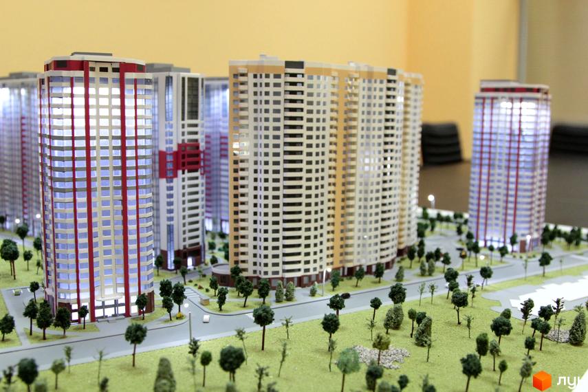 Макет житлового комплексу Позняки-4а з високими будинками, що мають скляні фасади з червоними та білими вертикальними елементами. Територія навколо моделі озеленена деревами та має дороги.