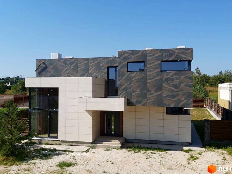 Будівля з бежевими та темно-сірими фасадами. Перед будинком пісок та зелень, на задньому плані паркан та дерева.