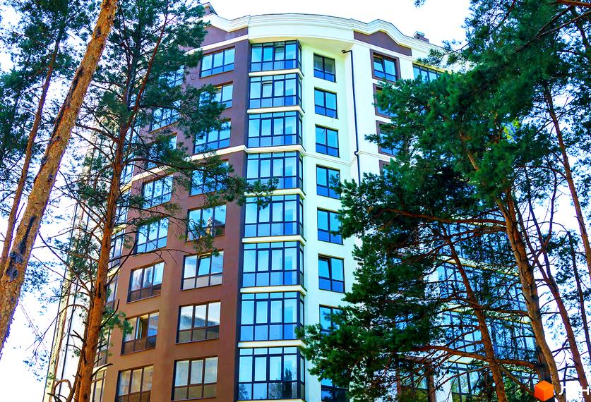 Зображення багатоповерхового будинку з великими вікнами. Житловий комплекс Лісова казка оточений високими деревами.