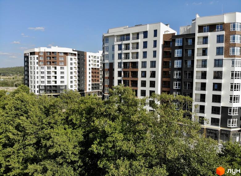 Житловий комплекс Синергія Сіті від Kvartal Group. Багатоповерхові будинки з білими, коричневими та чорними фасадами на тлі зелених дерев та блакитного неба.