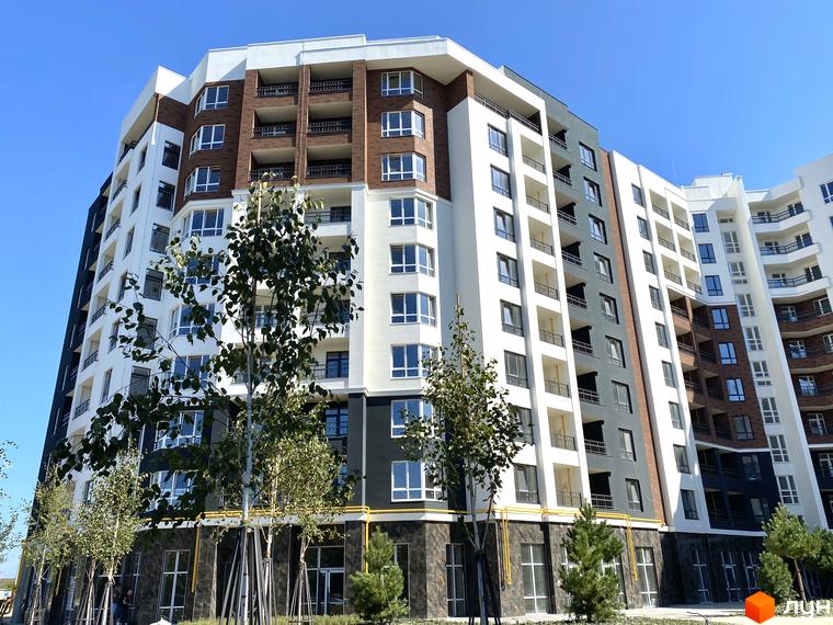 Зовнішній вигляд багатоквартирного будинку ЖК Синергія Сіті з білими, коричневими та сірими фасадами, балконами та великими вікнами. На передньому плані дерева та елементи прибудинкової території.