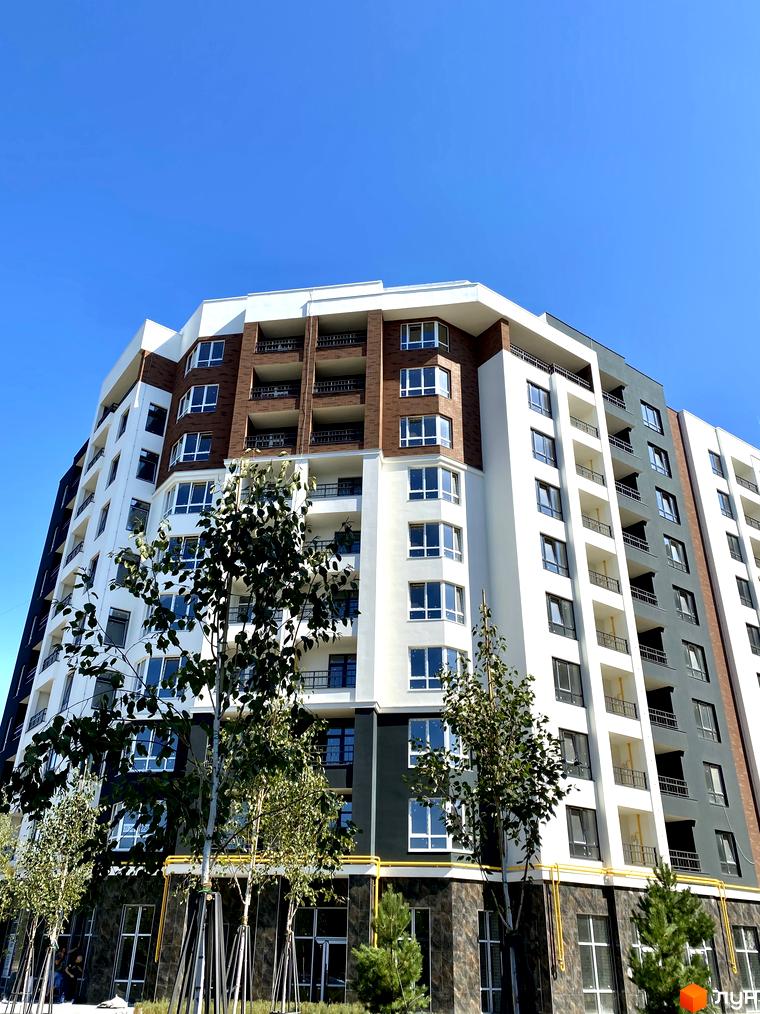 Фасад житлового комплексу Синергія Сіті з білими та коричневими елементами, балконами та великими вікнами. На передньому плані зелені дерева.