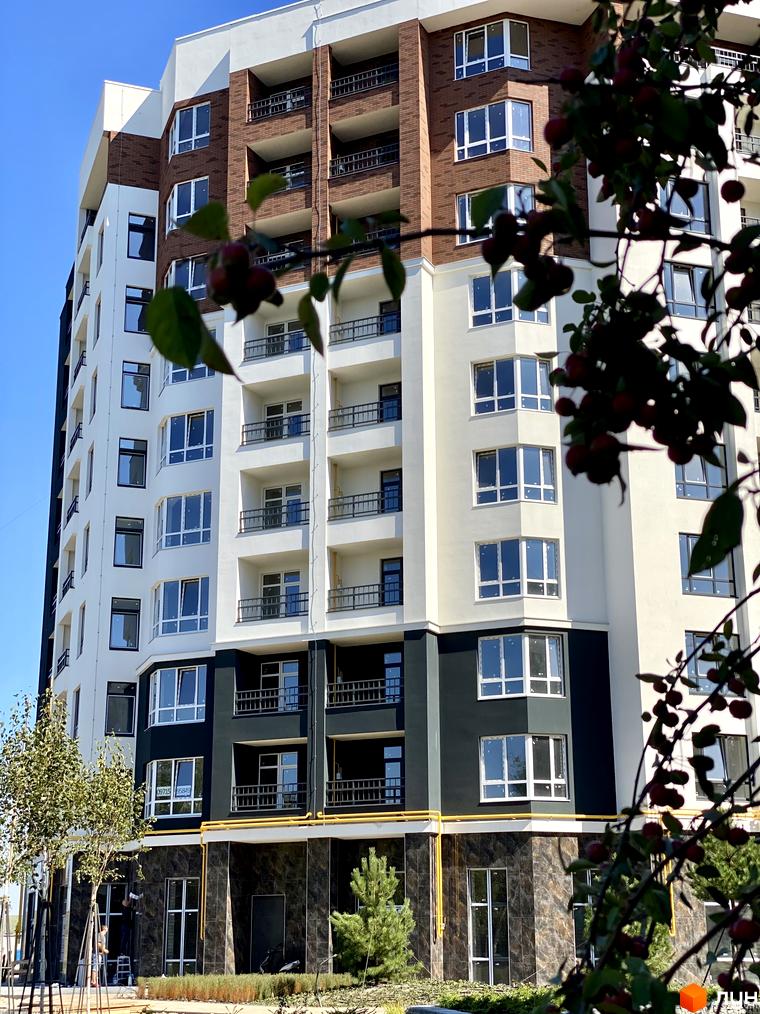 Будівельний проєкт ЖК Синергія Сіті від Kvartal Group з цегляним та білим фасадом, балконами та вікнами на тлі блакитного неба. На передньому плані гілка з червоними ягодами.