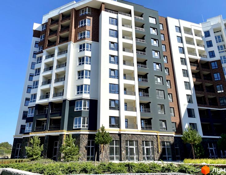 Зовнішній вигляд новобудови ЖК Синергія Сіті з фасадами з білої та сірої штукатурки, цегли та скляними балконами. Навколо комплексу зелені насадження та дерева.