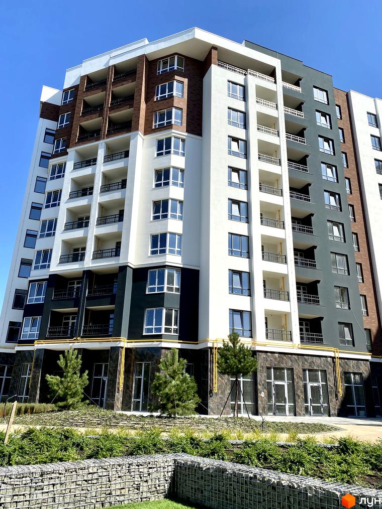 Сучасний житловий комплекс Синергія Сіті з білими та сірими фасадами, цегляними вставками та балконами. Прибудинкова територія з зеленими насадженнями та кам'яною огорожею.