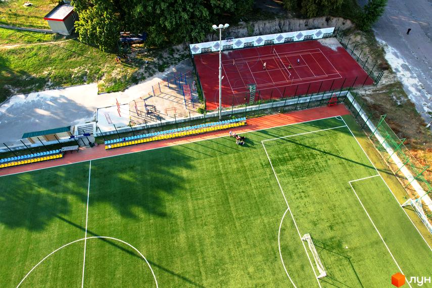 Котеджне містечко Панорама. Вид згори на зелене футбольне поле та червоний тенісний корт з людьми, що грають.