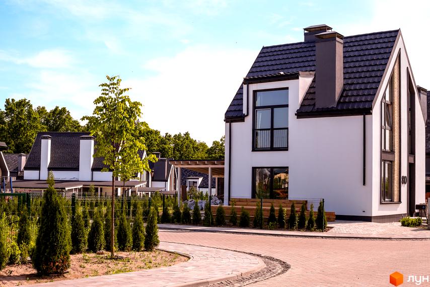 Котеджне містечко КМ VILLAGGO: білі фасади будинків з темними дахами та вікнами, зелені туї та доглянута прибудинкова територія з бруківкою.