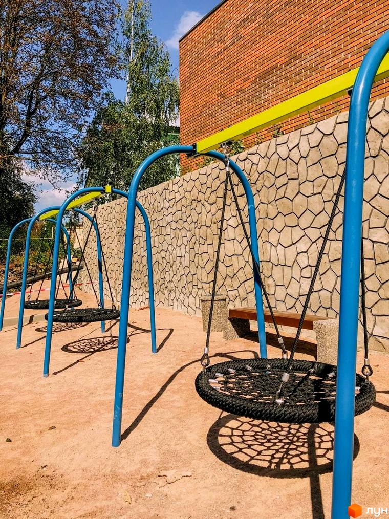 Дитячий майданчик з гойдалками у ЖК Преміум Парк. Територія біля будинку з лавкою та зеленню.