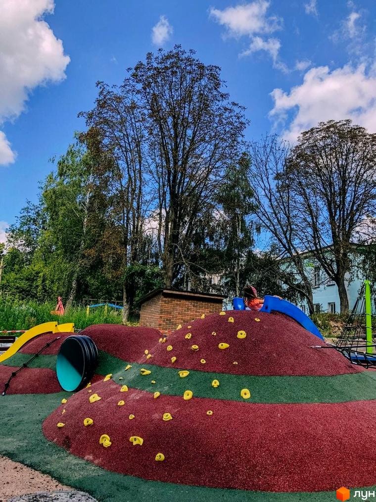 Дитячий майданчик в ЖК Преміум Парк з гіркою та скеледромом. Зелена територія з деревами та хмарним небом.