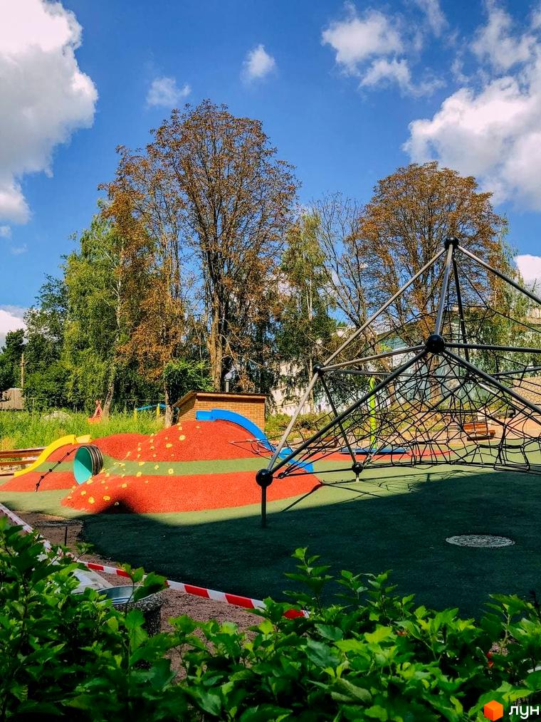 Дитячий майданчик у ЖК Преміум Парк з гірками, тунелями та мотузковим містечком. Зелена територія комплексу з деревами та хмарами на блакитному небі.