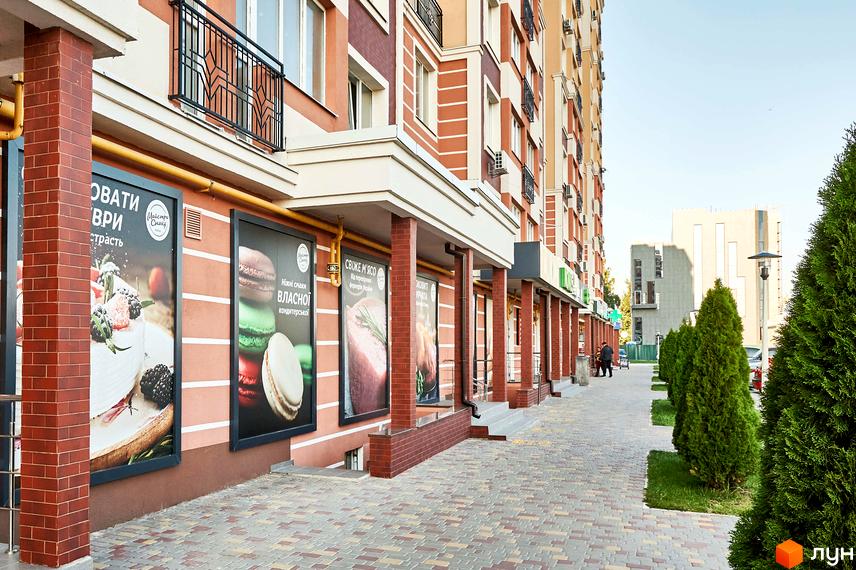 Фасад житлового комплексу Нові Теремки з цегляними колонами та комерційними вітринами. Поруч розташована вимощена алея з деревами.