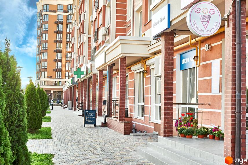 Житловий комплекс Нові Теремки. Фасад будинку з червоної цегли та бежевими вставками. Поруч зелені туї та бруківка.