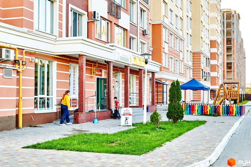 Фасад житлового комплексу з магазинами на першому поверсі. На прибудинковій території дитячий майданчик та зелені насадження.